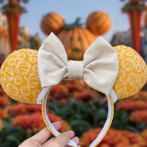 May include: A pair of yellow and white Minnie Mouse ears with a large white bow. The ears are covered in a pattern of small pumpkins.