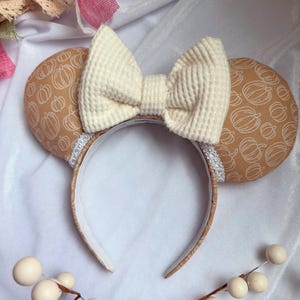 May include: A festive headband featuring two round ears with a pumpkin pattern in shades of brown and white. A large, textured cream-colored bow sits atop the headband. The headband is perfect for fall or Halloween.