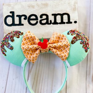 May include: Mint green mouse ears with a waffle cone bow and chocolate and sprinkles on top. The bow has a red Mickey Mouse icon in the center. The ears are on a headband.