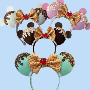 May include: Three pairs of ice cream themed Minnie Mouse ears. The ears are made of felt and feature a waffle cone, ice cream, sprinkles, and a cherry. The headband is white.