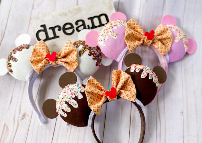 May include: Three pairs of ice cream themed Minnie Mouse ears. The ears are made of felt and feature a waffle cone bow, sprinkles, and a cherry. The ice cream flavors are chocolate, vanilla, and strawberry.