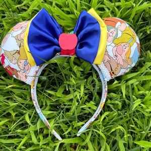 May include: A pair of blue and yellow fabric Minnie Mouse ears with a red bow. The ears are decorated with a pattern of the seven dwarfs from the Disney movie Snow White and the Seven Dwarfs.