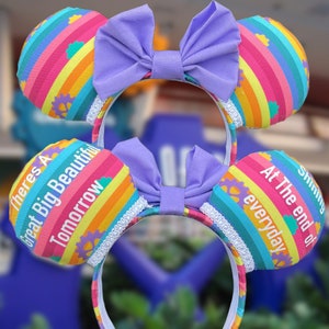 May include: Rainbow-striped fabric mouse ears with a purple bow. The left ear says "There's a Great Big Beautiful Tomorrow" and the right ear says "Shining At The End of Everyday".