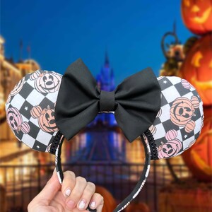 May include: Black and white checkered Minnie Mouse ears headband with a black bow. The ears feature a pink and white pumpkin design with a Minnie Mouse face.