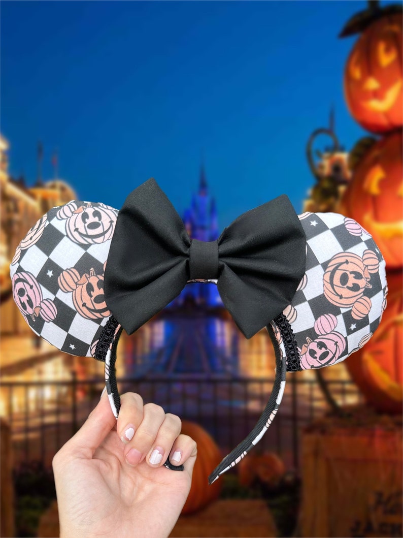 May include: Black and white checkered Minnie Mouse ears headband with a black bow. The ears feature a pink and white pumpkin design with a Minnie Mouse face.