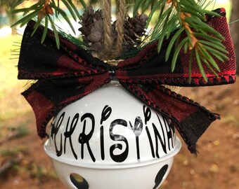 May include: White metal Christmas bell ornament with a black and red plaid bow and the name "Christina" written in black script.