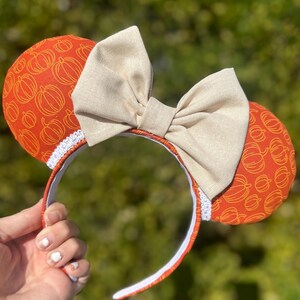 May include: Orange and white pumpkin-patterned fabric mouse ears with a large beige bow. The headband is white with a white lace trim.
