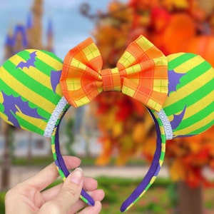 May include: Green and yellow striped Minnie Mouse ears with a purple bat design and a plaid orange and yellow bow.
