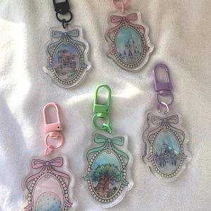 May include: Five colorful keychains with different Disney park images. Each keychain features a clear oval charm with a white pearl border, a pink bow, and a metal clasp. The charms feature images of Cinderella's Castle, the Tree of Life, and the Magic Kingdom.