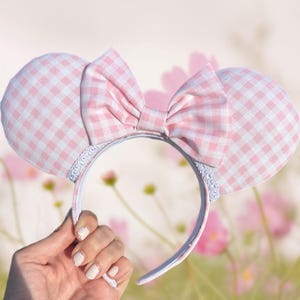 May include: Pink and white gingham pattern mouse ear headband with a matching bow. The ears and bow are made of a soft fabric. The headband is held by a hand against a blurred floral background.