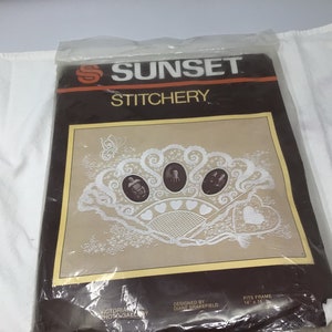 May include: A white embroidery kit with a lace design featuring three oval-shaped photo frames. The kit is packaged in a clear plastic bag with a brown and white label that reads "Sunset Stitchery Victorian Photo Gallery Designed by Diane Brakefield Fits Frame 14" x 18"