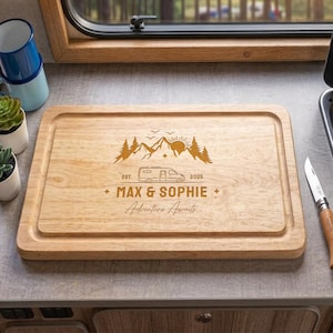 May include: A rectangular wooden chopping board with a juice groove and a laser-engraved design featuring mountains, a camper van, the text EST. 2025, the names MAX & SOPHIE, and the phrase Adventure Awaits centred on the light brown wood surface.