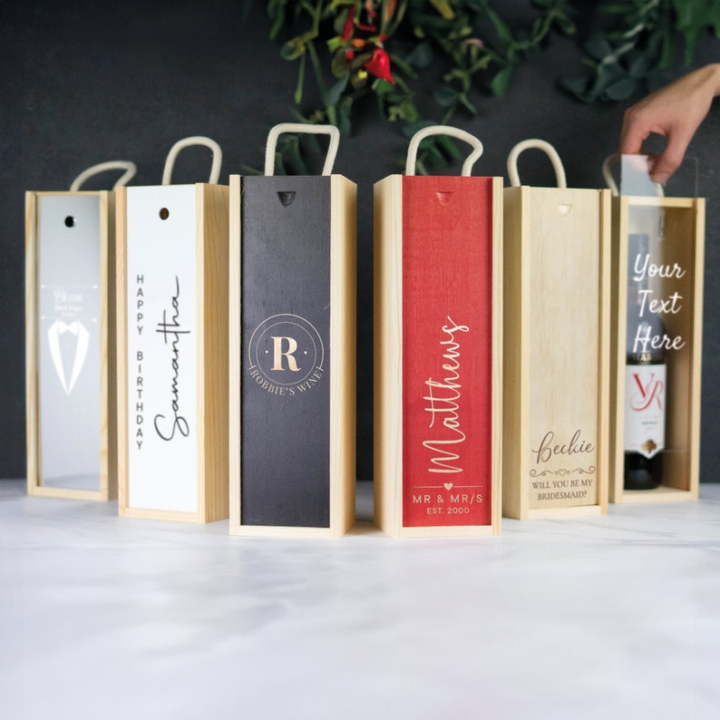May include: Five personalized wooden wine boxes with rope handles. The boxes are in various colors, including gray, natural wood, black, red, and natural wood. The boxes are engraved with different messages, including "Happy Birthday Samantha", "R Robbie's Wine", "Matthews MR & MRS EST. 2000", "Beckie Will You Be My Bridesmaid", and "Your Text Here".