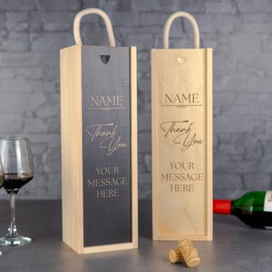 May include: Two wooden wine boxes with rope handles. One box has a dark grey front panel, the other is natural wood. Both boxes are engraved with "NAME", "Thank You", and "YOUR MESSAGE HERE". A wine glass and corks are in the foreground.