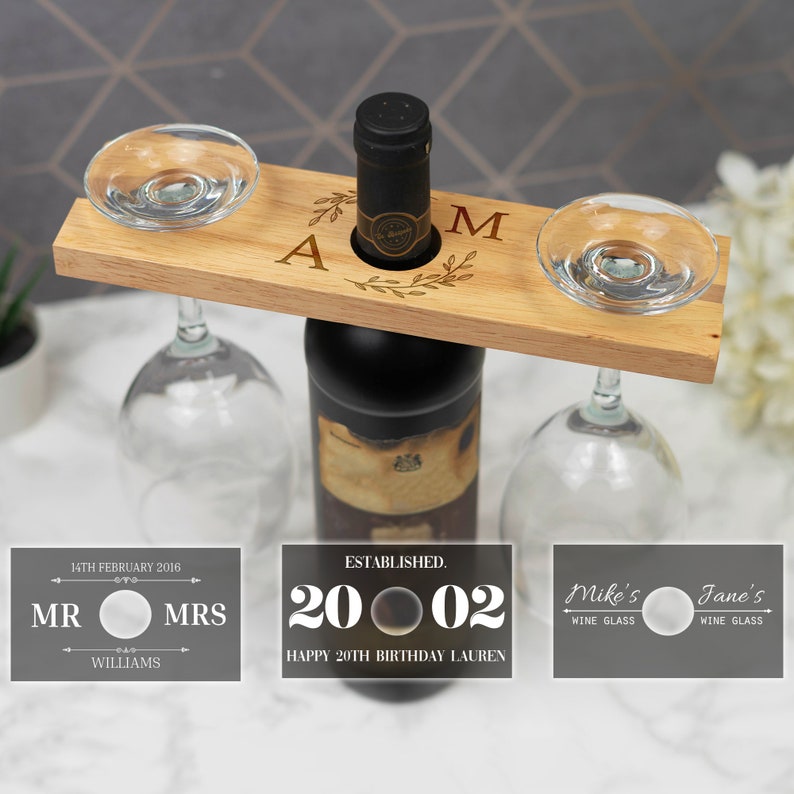 May include: Wooden wine bottle holder with two glass holders. The holder is engraved with the letters "A" and "M" and a floral design. There are three labels on the table below the holder. The first label says "14th February 2016 MR MRS WILLIAMS". The second label says "ESTABLISHED. 20 02 HAPPY 20TH BIRTHDAY LAUREN". The third label says "Mike's WINE GLASS Jane's WINE GLASS".