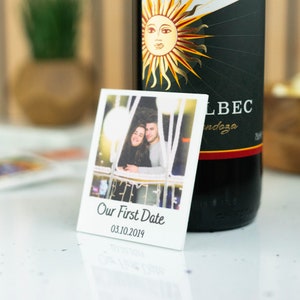 May include: A white square photo with a couple posing in front of a lit-up background. The photo has the text "Our First Date" and the date "03.10.2019" printed below the image.