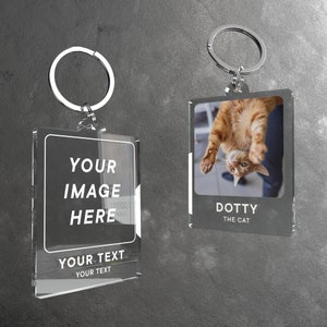 Puede incluir: Dos marcos de fotos de llavero de acr&iacute;lico transparente con llaveros plateados. El marco de la izquierda tiene el texto "YOUR IMAGE HERE" y "YOUR TEXT YOUR TEXT". El marco de la derecha tiene una foto de un gato boca abajo con el texto "DOTTY THE CAT".