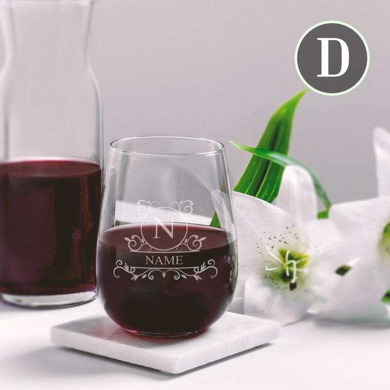 May include: A stemless wine glass with a personalised monogram design. The glass is filled with red wine and sits on a white coaster. The monogram features the letter "N" inside a circle with the word "NAME" below it.