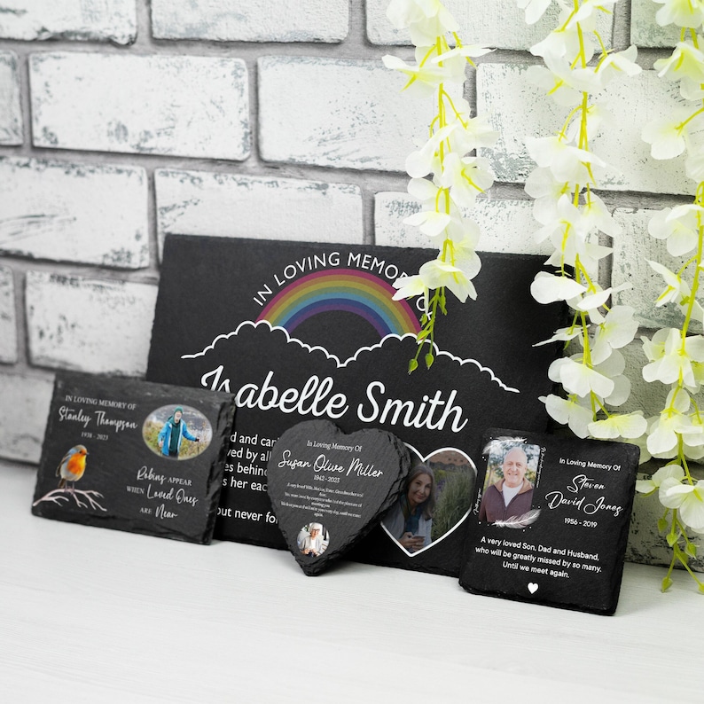 K&ouml;nnte beinhalten: Ein Set aus vier schwarzen Schiefer-Gedenktafeln mit Regenbogen-Designs und personalisierten Botschaften. Jede Tafel zeigt ein Foto und eine Gedenknachricht. Die Tafeln sind auf einem Ziegelhintergrund mit wei&szlig;en Blumen angeordnet.