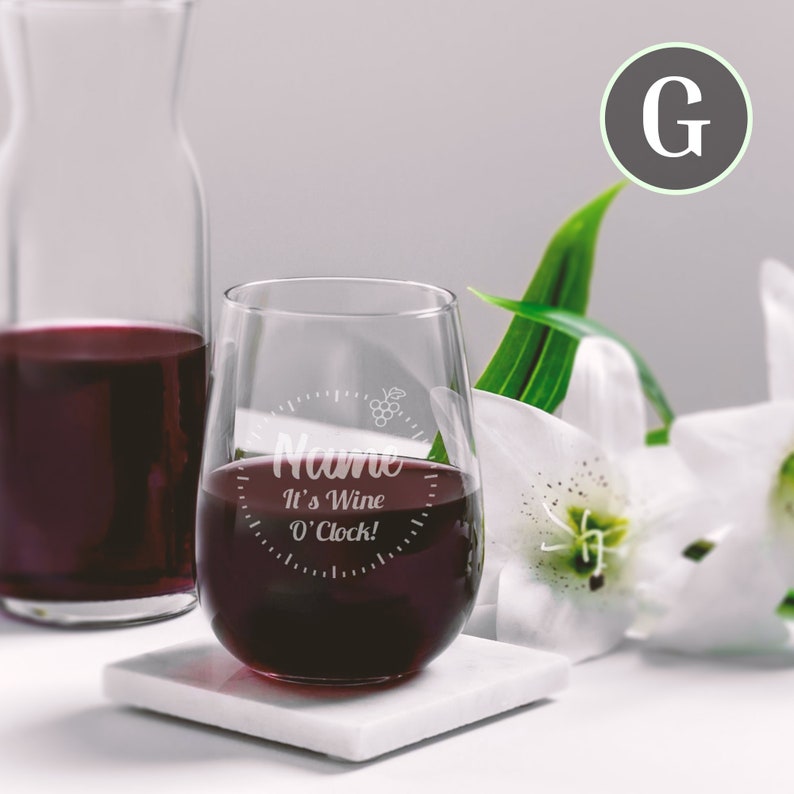 May include: A clear stemless wine glass with the text "Name It's Wine O'Clock!" etched on the side. The glass is filled with red wine and is sitting on a white coaster.