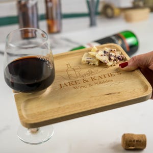 Può includere: Un vassoio di legno con la scritta "Jake & Katie's Wine & Nibbles" contiene formaggio ed è tenuto da una persona. Un bicchiere da vino senza stelo con vino rosso è sul vassoio. Una bottiglia di vino è sullo sfondo.
