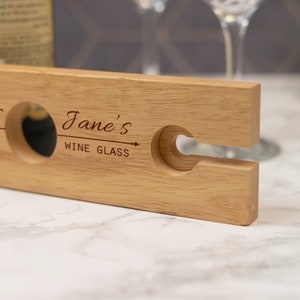 May include: A wooden wine glass holder with a cutout for a wine glass and a bottle. The holder is engraved with the text "Jane's Wine Glass".