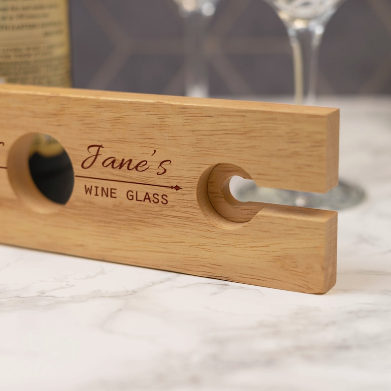 May include: A wooden wine glass holder with a cutout for a wine glass and a bottle. The holder is engraved with the text "Jane's Wine Glass".