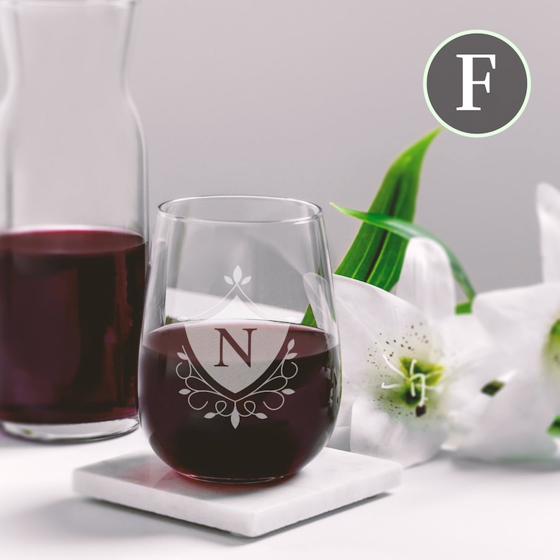 May include: A stemless wine glass with a personalised monogram design. The glass is etched with a shield and the letter "N" inside. The glass is filled with red wine and is sitting on a white coaster.