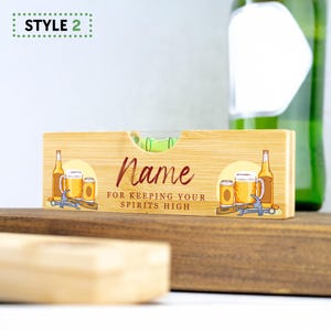 K&ouml;nnte beinhalten: Ein Holz-Wasserwaage mit einer gr&uuml;nen Libelle und einem bedruckten Design von Bierflaschen und Bechern. Die Wasserwaage tr&auml;gt den Text "Name for keeping your spirits high".