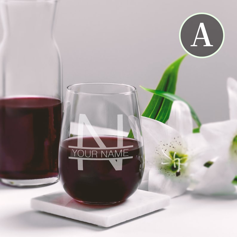 May include: A stemless wine glass with the letter "N" etched on the front. The glass is filled with red wine and has the text "YOUR NAME" etched below the letter. The glass is sitting on a white coaster.
