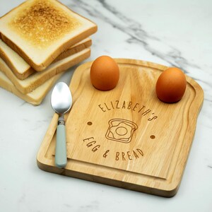 May include: A personalised wooden breakfast board with two egg holders, a spoon, and a stack of toast. The board is engraved with the text "Elizabeth's Egg & Bread" and a stylised image of a fried egg.