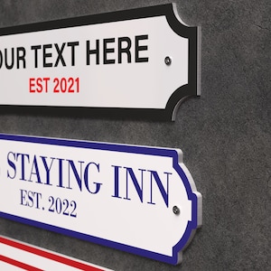 May include: White rectangular signs with decorative edges and text. One sign reads "YOUR TEXT HERE" and "EST 2021" in red. Another sign says "STAYING INN" and "EST. 2022" in blue. The signs are mounted on a grey wall.