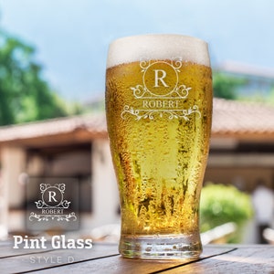 Op de afbeelding: Een gepersonaliseerd pintglas met een mat ontwerp. Het glas heeft een gegraveerde initialen "R" in een cirkel met een decoratieve versiering en de naam "Robert" eronder. Het glas is gevuld met een gouden drank.