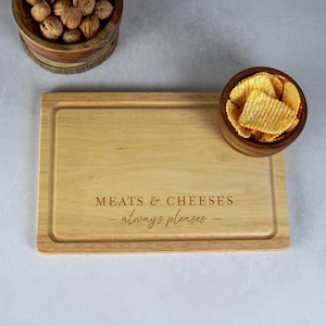 May include: A rectangular wooden serving board with the engraved text "MEATS & CHEESES - always pleases -". The board is surrounded by a small rim. A small wooden bowl of potato chips and a bowl of walnuts are also present.