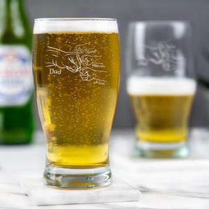 May include: A pint glass filled with golden beer and a foamy head. The glass features an etched design of fists with the word "Dad" and the names "Oliver," "Mason," and "Jessie." Another glass is visible in the background.