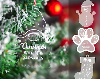 May include: Four clear acrylic Christmas ornaments with a white design. The ornaments are shaped like a gnome, a snowman, a paw print, and a stocking. The gnome ornament has the text "Christmas with the Surname's". The snowman ornament has the text "Name". The paw print ornament has the text "Name". The stocking ornament has the text "Name".