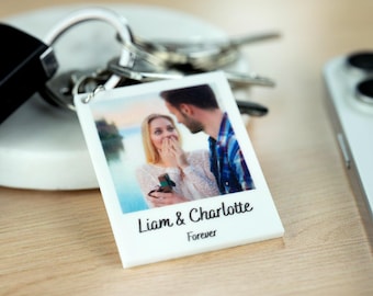 Könnte beinhalten: Ein weißer Schlüsselanhänger mit einem Foto eines Paares und dem Text "Liam & Charlotte Forever". Der Schlüsselanhänger ist an einem Schlüsselbund befestigt. Im Hintergrund ist ein weißes Telefon zu sehen.
