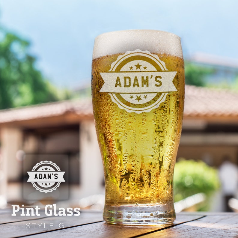 Op de afbeelding: Een bevroren pintglas met een gepersonaliseerd etiket met de tekst "Adam's" in een gouden cirkel met sterren. Het glas is gevuld met een goudbruin bier.