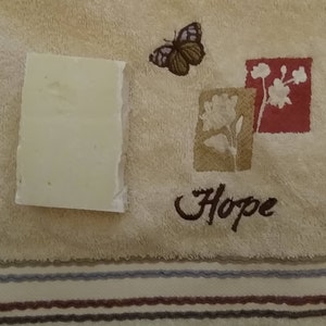 May include: A rectangular bar of pale green soap rests on a beige towel. The towel features embroidered floral designs in red and tan, a butterfly, and the word "Hope".