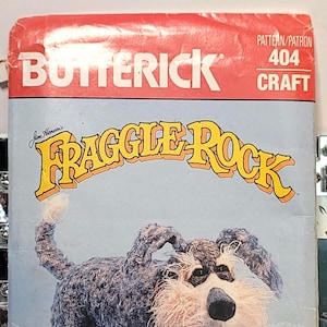 May include: A Butterick 404 Craft sewing pattern envelope for a Jim Henson's Fraggle Rock Sprocket dog plush. The cover features a graphic of a shaggy, dark gray and white dog with a pink tongue, set against a light blue background with yellow text.