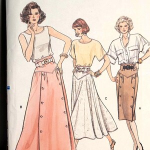 Puede incluir: Sobre de patrón de costura Vogue Patterns 9851 para tallas 12-14-16. La ilustración muestra tres conjuntos: una falda rosa con botones y top sin mangas, una falda clara con top de manga corta y una falda marrón con camisa de botones.