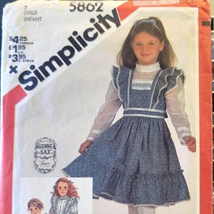 Może przedstawiać: Koperta z wykrojem krawieckim Simplicity 5862 przedstawiająca niebieską sukienkę w kwiatowy wzór z białymi koronkowymi wykończeniami, wysokim kołnierzem i przezroczystymi długimi rękawami. Okładka zawiera zdjęcie dziecka oraz dwie ilustracje.