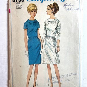 Op de afbeelding: Een vintage Simplicity 6799 naaipatroon-envelop voor een jurk in maat 14, buste 34. De illustratie toont twee variaties van een jurk: een in effen blauw met korte mouwen en een riem, en een in witte stof met patroon en driekwartmouwen.
