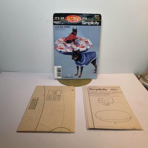 May include: A Simplicity 3664 sewing pattern envelope featuring a dog wearing a red coat and a blue coat with white trim, alongside two tan paper pattern instruction sheets. The envelope displays the text IT'S SO easy IT'S Simplicity 3664.