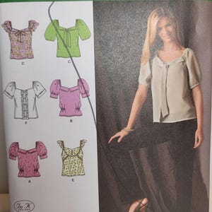 Könnte beinhalten: Simplicity Schnittmuster 2697, Größe D5 4, 6, 8, 10, 12. Die Hülle zeigt Illustrationen von sechs verschiedenen Blusenmodellen mit unterschiedlichen Ärmellängen, Ausschnitten und Mustern sowie ein Foto eines Modells in einer beigen Bluse.