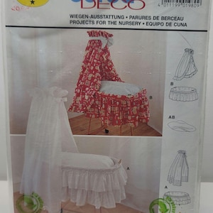 May include: A Burda Deco sewing pattern package for nursery projects, labeled 1980. The packaging displays two cradle designs: one with a red patterned fabric canopy and skirt, and another with a white sheer canopy and ruffled white skirt.
