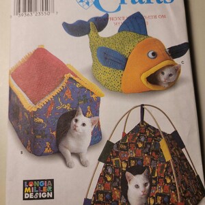May include: A Simplicity Crafts sewing pattern envelope 9004 featuring illustrations of three pet beds: a fish-shaped bed, a house-shaped bed with a cat print, and a tent-style bed with a cat print. Each bed contains a white cat illustration.