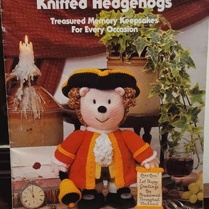 Könnte beinhalten: Buchcover "Jean Greenhowe's Knitted Hedgehogs" mit einem gestrickten Igel in roter und gelber Kleidung. Der Text "Treasured Memory Keepsakes For Every Occasion" ist zu sehen. Ein kleines Schild lautet "Oyez Oyez! Let Happy Greetings Be Proclaimed Throughout The Land."