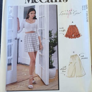 May include: McCall's sewing pattern M8221, featuring a woman wearing a white top and a black and white plaid skirt. The pattern includes illustrations for making shorts with ruffles and side pockets. The text "Learn to Sew" is visible.