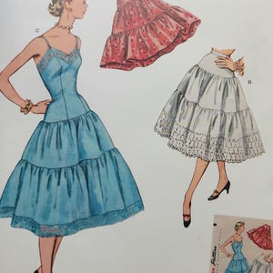 Puede incluir: Patrón de costura Simplicity vintage de los años 1950. La ilustración presenta un vestido azul con falda escalonada, una enagua roja y una enagua blanca con ribete de encaje. El patrón incluye información de tallas.